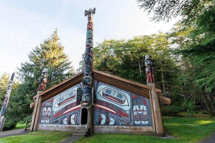A painted clan house with a tall totem pole at its door, spruce all around