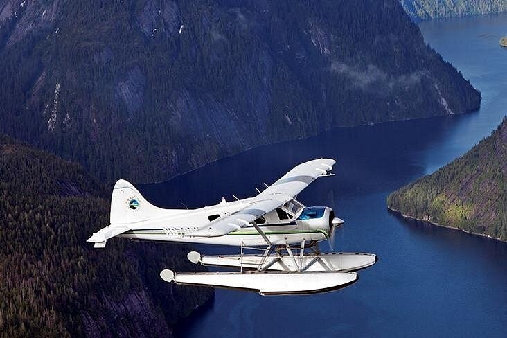 A white floatplane flying low along a steep-sided fjord
