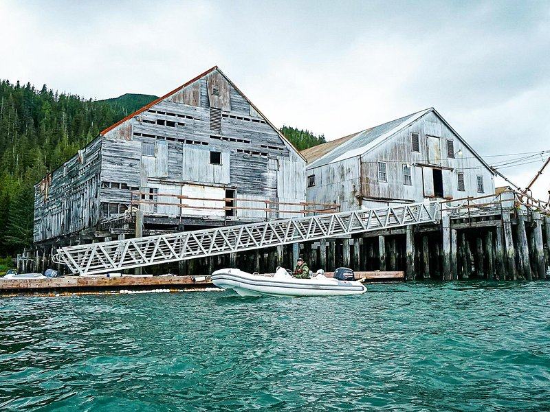 A weathered wooden cannery building on pilings over green water